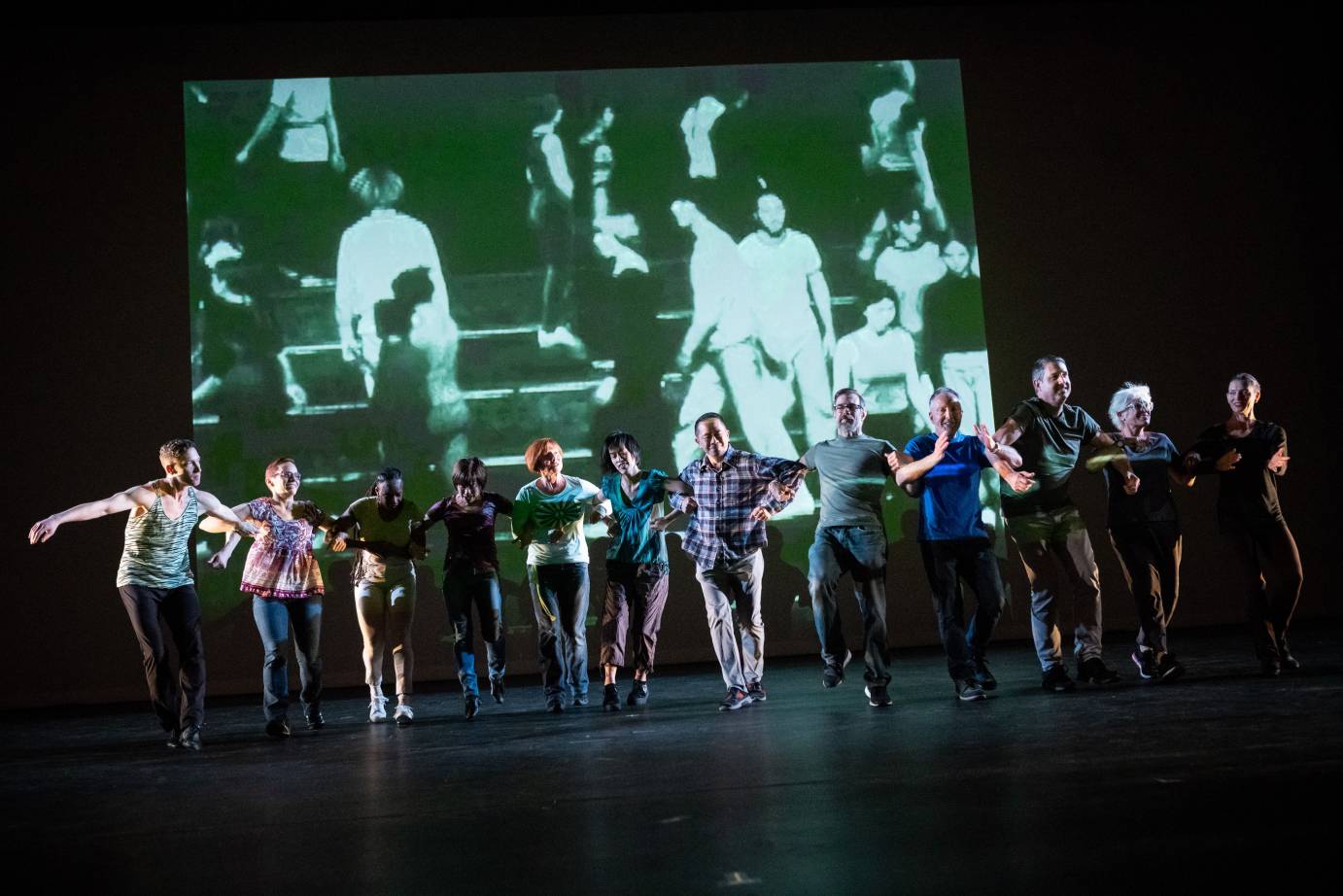 A grainy archival video of Twyla Tharp dancers is projected on stage. In front of it, a group of dancers and volunteers stand in a straight line with their arms linked.